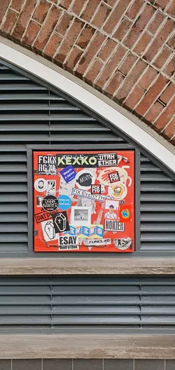 A red surface covered in various stickers, including designs with text and images. Some stickers appear to be from artists or brands. The stickers are densely packed, creating a collage-like effect. The surface is mounted on a wall with brickwork above and slats below. The stickers feature a range of styles, from hand-drawn to professionally printed.