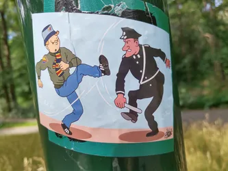 The sticker shows a man in a jacket and jeans kicking a policeman who is holding a baton. The style is reminiscent of comic strips or cartoons.  The colors are mainly blue, brown and black on a light blue background. The sticker appears to be applied to a green pole.