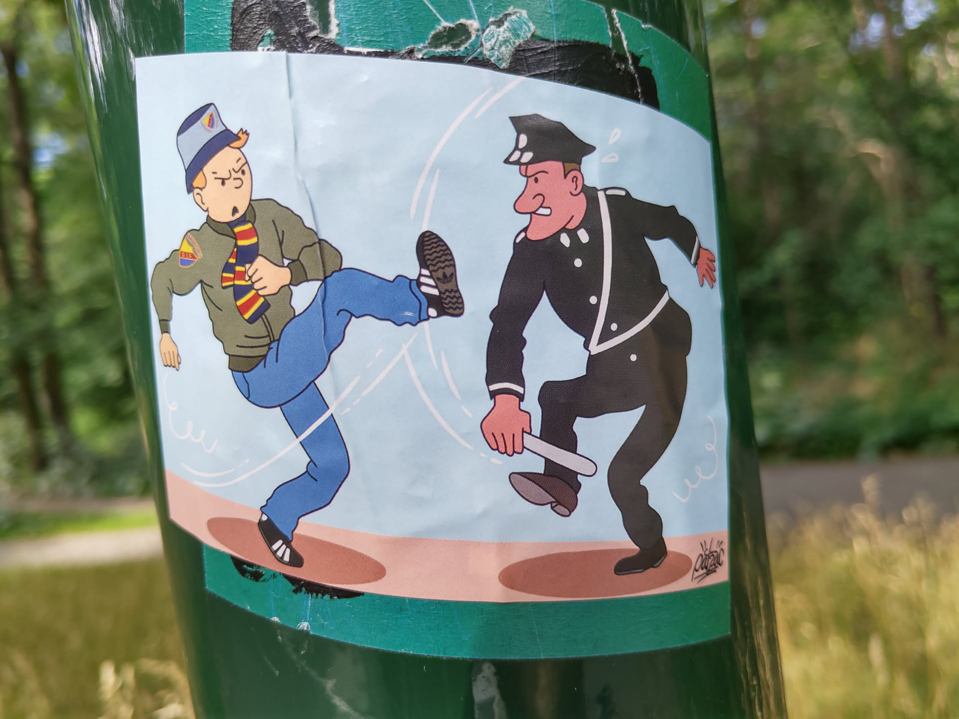 The sticker shows a man in a jacket and jeans kicking a policeman who is holding a baton. The style is reminiscent of comic strips or cartoons.  The colors are mainly blue, brown and black on a light blue background. The sticker appears to be applied to a green pole.