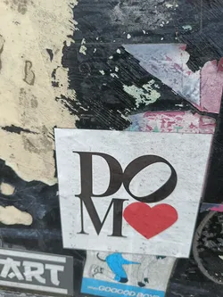 A minimalist sticker with the text 'DO MORE' and a red heart. The sticker is affixed to a grunge background with other stickers and markings.