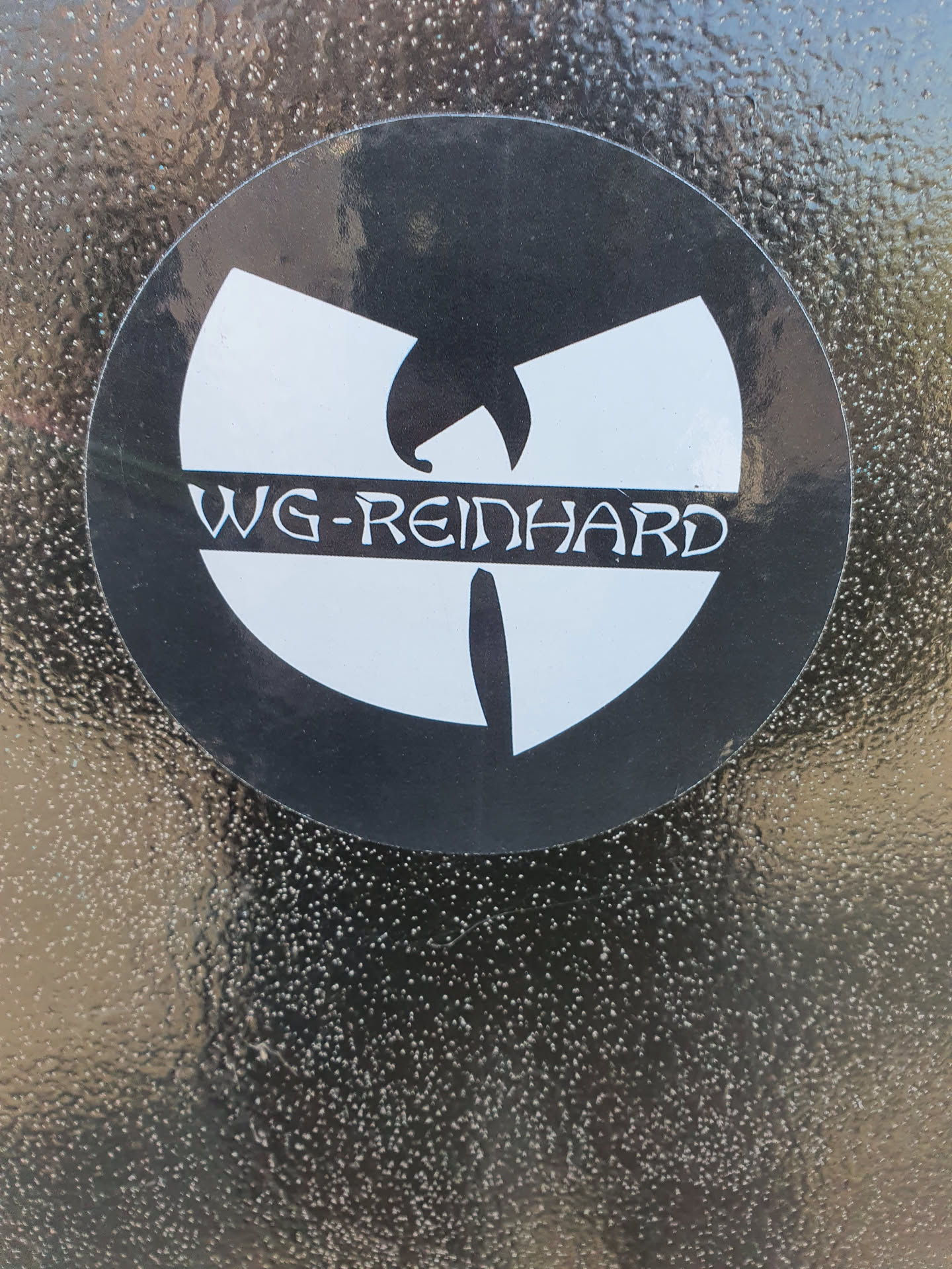 A round sticker featuring a stylized Wu-Tang Clan logo with "WG-REINHARD" written underneath. The sticker is black and white and is applied to a textured dark surface. 