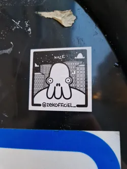Black and white illustration of a character resembling an octopus with large eyes, set against a cityscape backdrop. The sticker features the artist's Instagram handle and the word "KOZE" at the top.