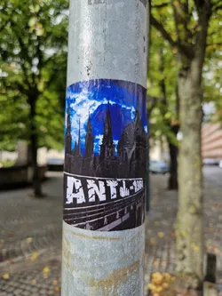 A sticker featuring a stylized image of a city skyline under a blue sky with clouds, and the text 'ANTL-1964' at the bottom. The style is reminiscent of vintage posters or prints.