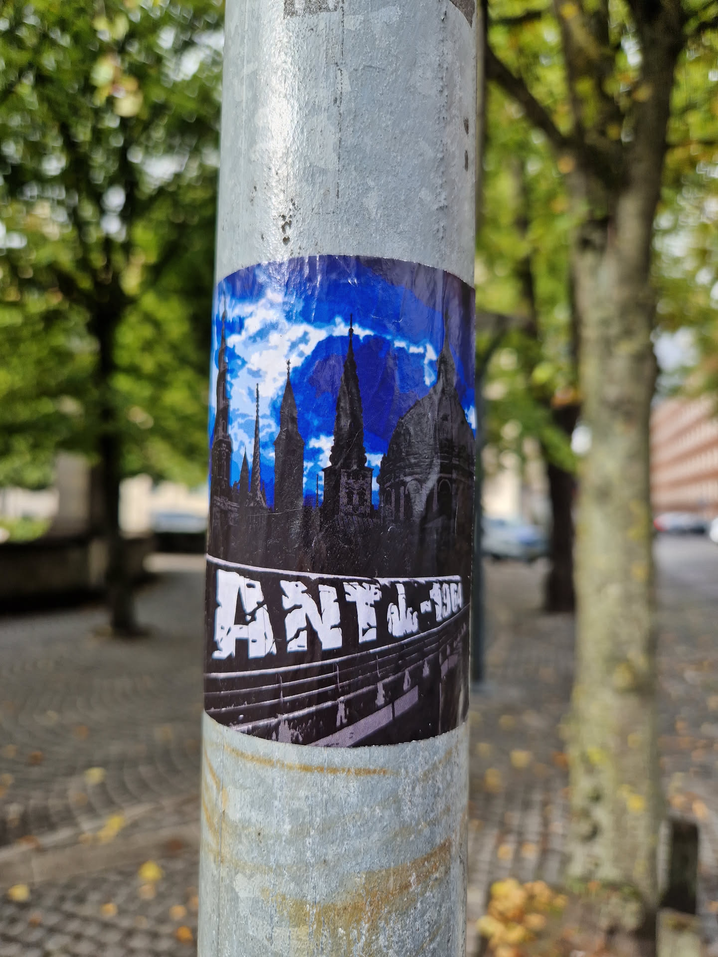 A sticker featuring a stylized image of a city skyline under a blue sky with clouds, and the text 'ANTL-1964' at the bottom. The style is reminiscent of vintage posters or prints.