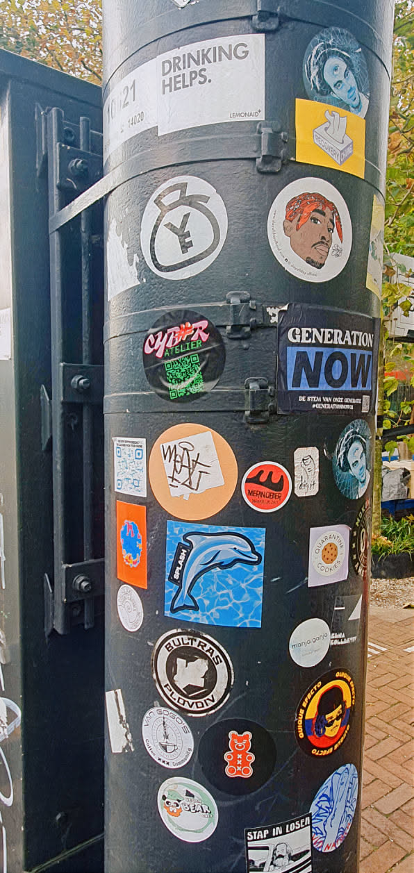 This image shows a black post densely covered with a variety of stickers. Centrally located is a blue sticker featuring a dolphin jumping out of the water with the word "SPLASH" printed on it. Other stickers display diverse designs, including text, logos, and illustrations, contributing to the post's eclectic appearance.