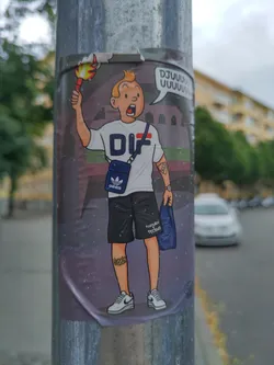 A sticker featuring a stylized illustration of Tintin, the famous comic book character. He is depicted wearing Adidas clothing and carrying a shopping bag. He is holding a flaming torch. The background shows a stadium. The sticker has a slightly damaged upper edge.