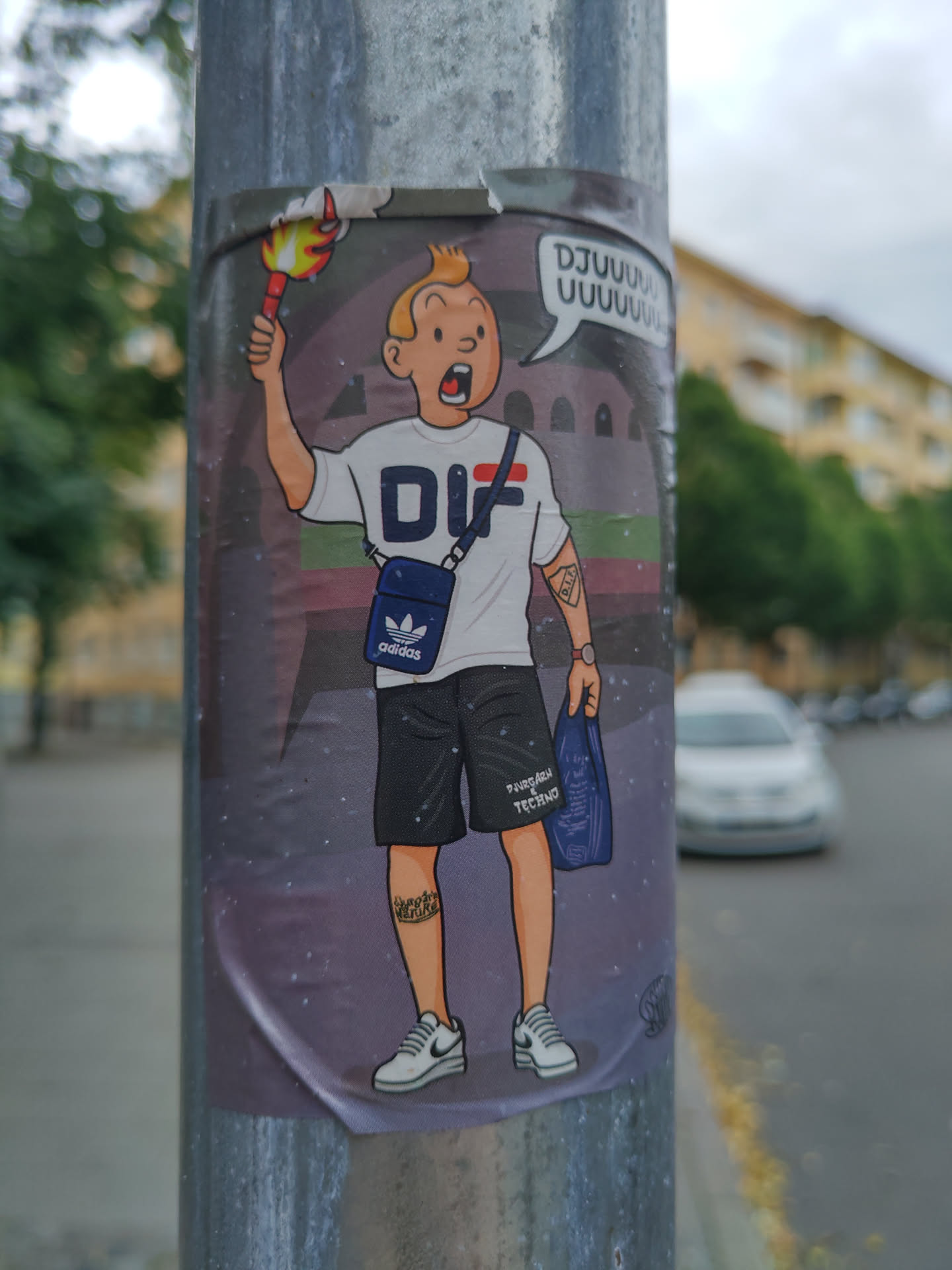 A sticker featuring a stylized illustration of Tintin, the famous comic book character. He is depicted wearing Adidas clothing and carrying a shopping bag. He is holding a flaming torch. The background shows a stadium. The sticker has a slightly damaged upper edge.