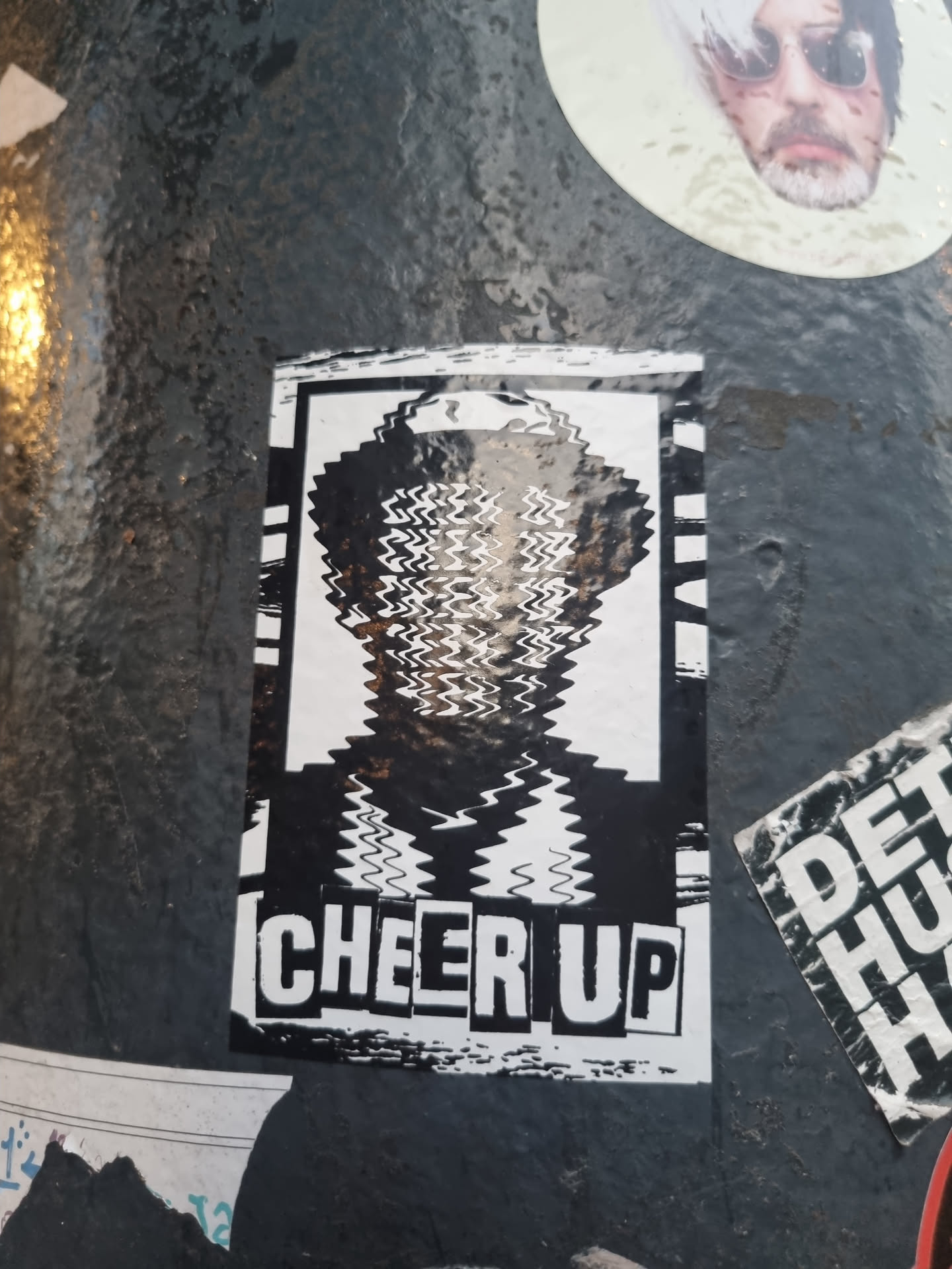 Black and white sticker with a distorted image of a person and the words "CHEER UP" at the bottom. The style is reminiscent of glitch art or digital distortion.