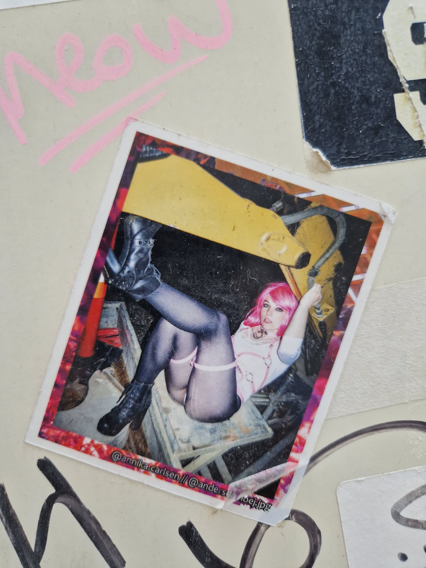 A sticker featuring a photo of a girl with pink hair sitting on a chair, with an excavator in the background. The sticker has a grunge, urban aesthetic and includes the artist's social media handles.