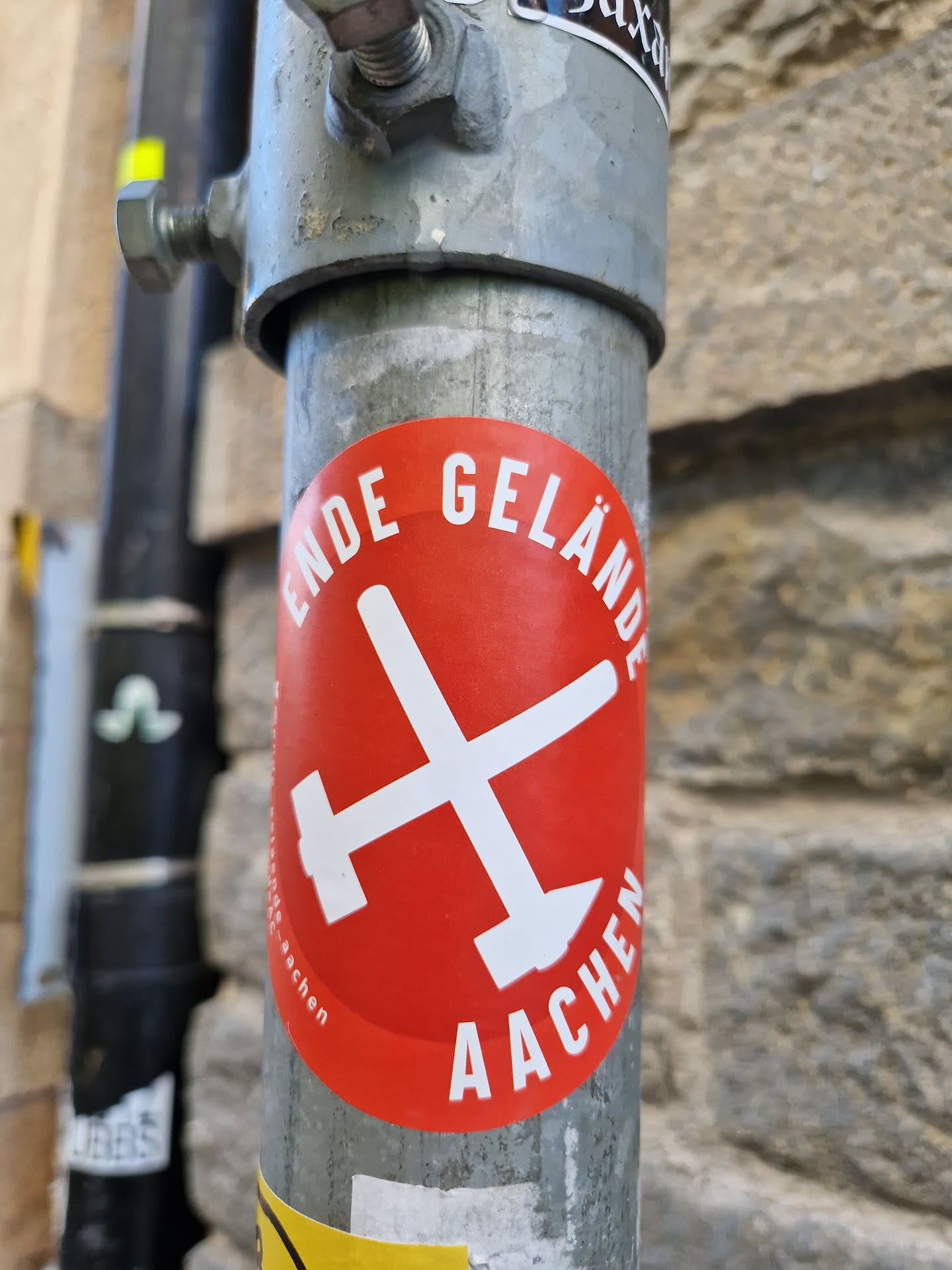 A red circular sticker with a white X formed by two crossed hammers. The text "Ende Gelände Aachen" surrounds the symbol.  The sticker is affixed to a metal pole.