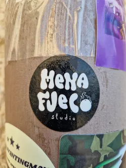 A black and white round sticker with the text "Mena Fuego studio" in a stylized font. It features a small flame logo next to the text.