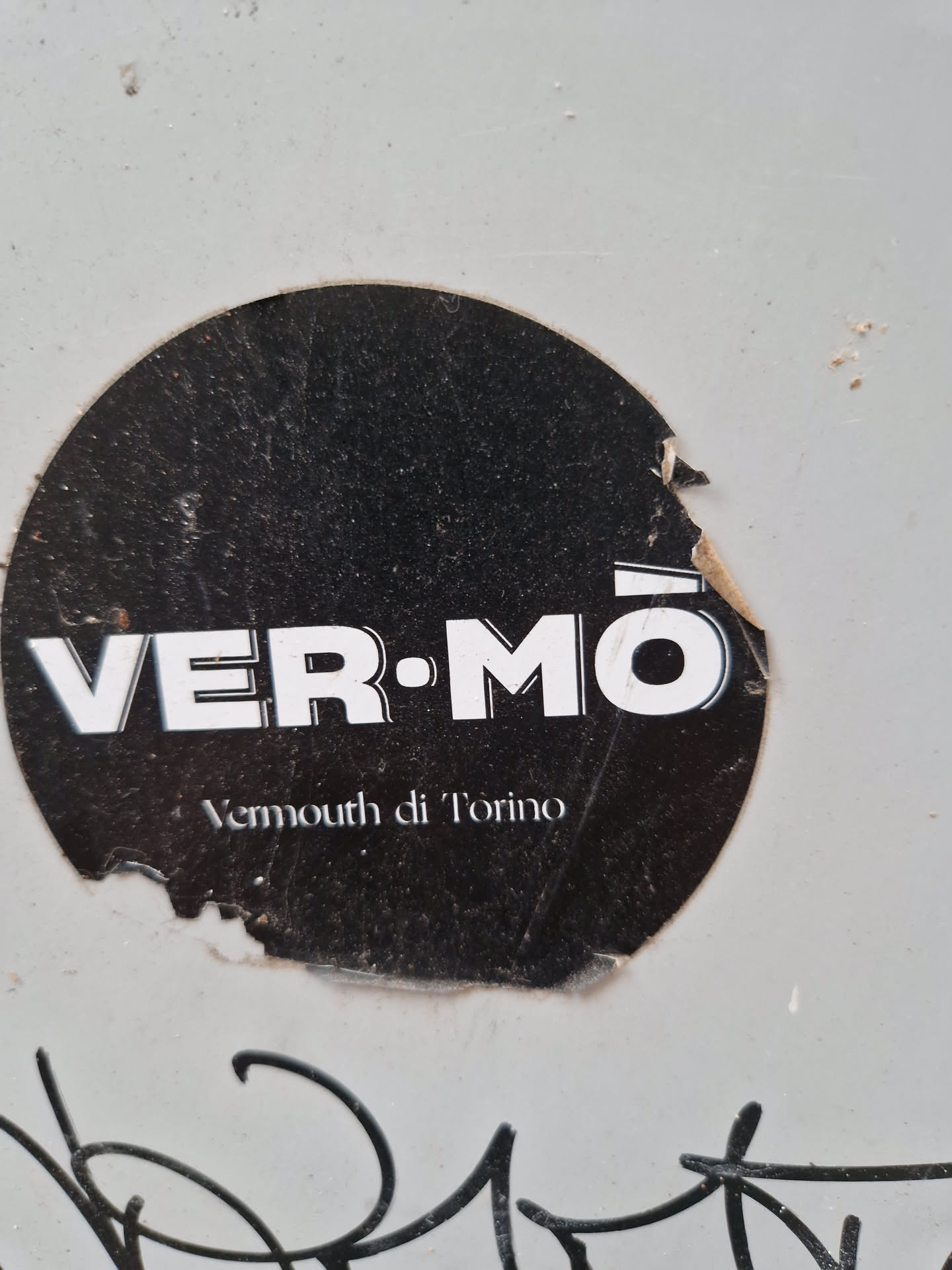 A round black and white sticker with the text VERMŌ and Vermouth di Torino. The sticker is slightly damaged.