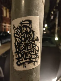 A black and white sticker with graffiti-style lettering. The text says "Old School Never Dies". The sticker is attached to a metal pole at night.