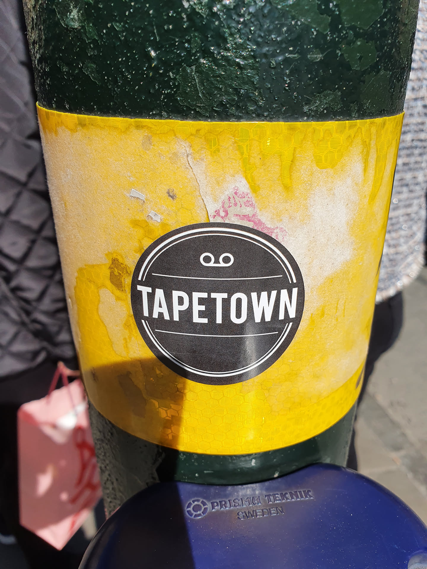 A worn yellow sticker with a black circular logo in the center. The logo says TAPETOWN in a sans-serif font with two small circles above the text. The sticker is affixed to a dark green pole.  The yellow sticker shows significant wear and tear.