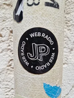 A black circular sticker with the letters JP in a stylized font. The text 'WEB RADIO' is written around the circle in a sans-serif font.