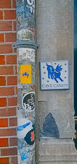 The photo shows a yellow sticker affixed to a weathered metal pipe amidst other urban stickers and graffiti. The sticker displays an abstract design in blue and orange. To the right, mounted on a stone wall, is a traditional 'Cave Canem' sign depicting a dog. The scene suggests a blend of contemporary urban culture and classical motifs.