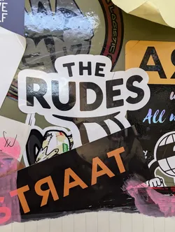 A black and white sticker with the text "THE RUDES" in a bold, sans-serif font. The sticker has a glossy finish.