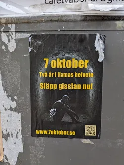 The sticker features a dark, gritty image of what appears to be a tunnel or confined space. In the center, a silhouetted figure is seated, appearing distressed and possibly chained. The text is in bold yellow lettering. The top line reads '7 oktober', followed by 'Två år i Hamas helvete' (Two years in Hamas hell), and 'Släpp gisslan nu!' (Release the hostages now!). A QR code is present in the bottom right corner, and the website 'www.7oktober.se' is displayed below the main text.