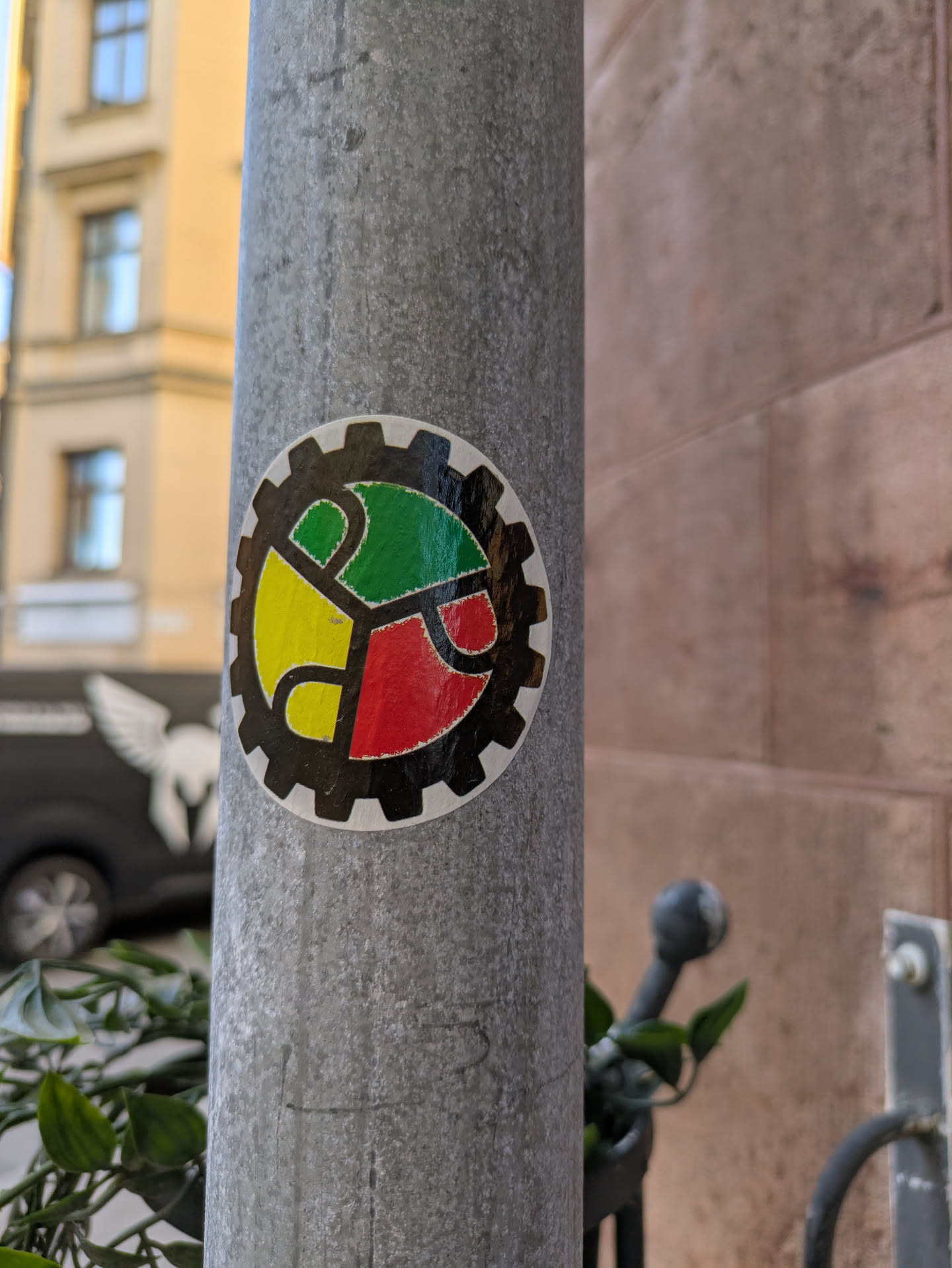 A sticker on a gray pole, featuring a gear-shaped design. The gear is divided into three sections colored yellow, green, and red, each containing a stylized 'R'.