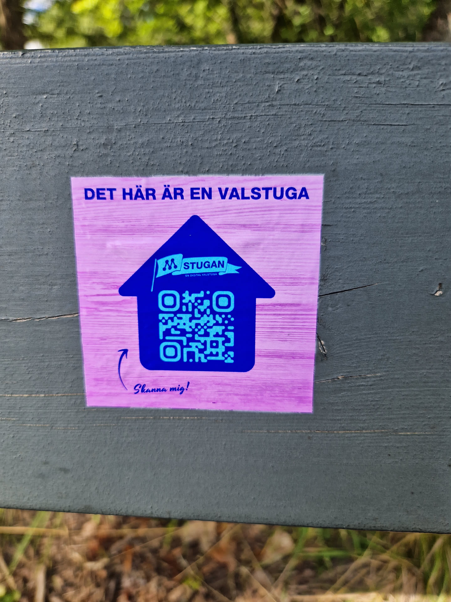 Purple and blue house-shaped sticker with a QR code.  The text at the top says "DET HÄR ÄR EN VALSTUGA" (This is a polling station). Inside the house shape is the logo for "M Stugan" and a QR code. At the bottom, it says "Scanna mig!" (Scan me!).
