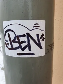 A square white sticker with dark purple graffiti that says BEN. The sticker is on a grey pole.