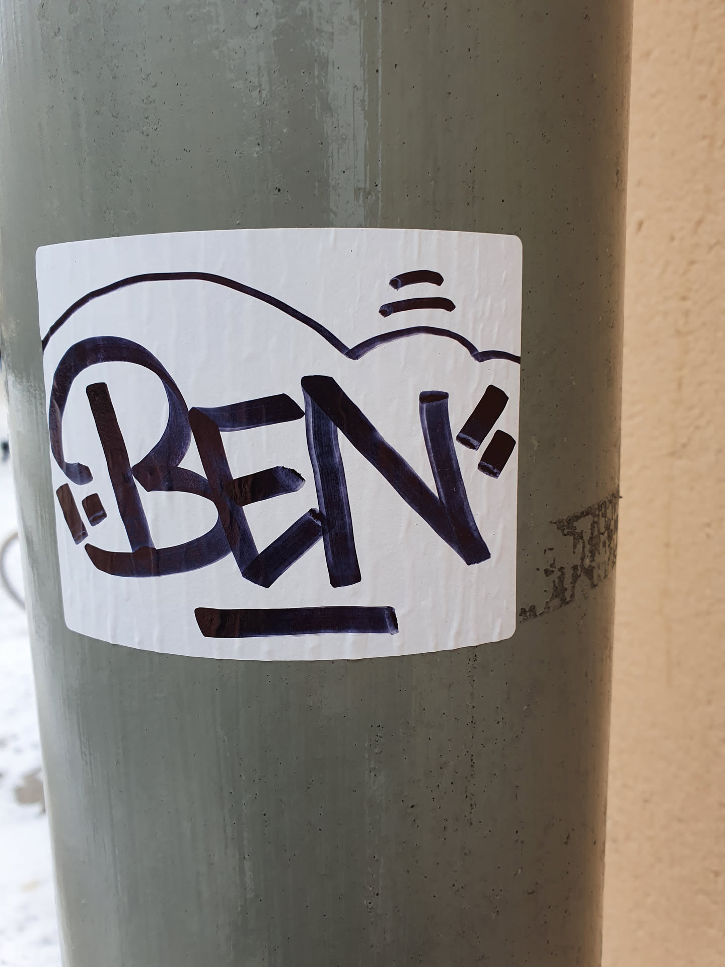 A square white sticker with dark purple graffiti that says BEN. The sticker is on a grey pole.