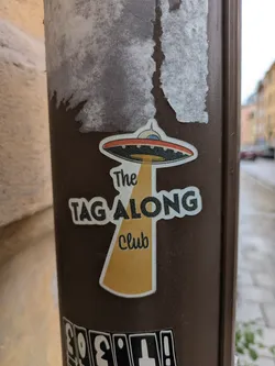 This sticker features an illustration of a flying saucer with a beam of light shining down from it. Underneath the beam of light are the words "The Tag Along Club". The sticker is on a brown pole.