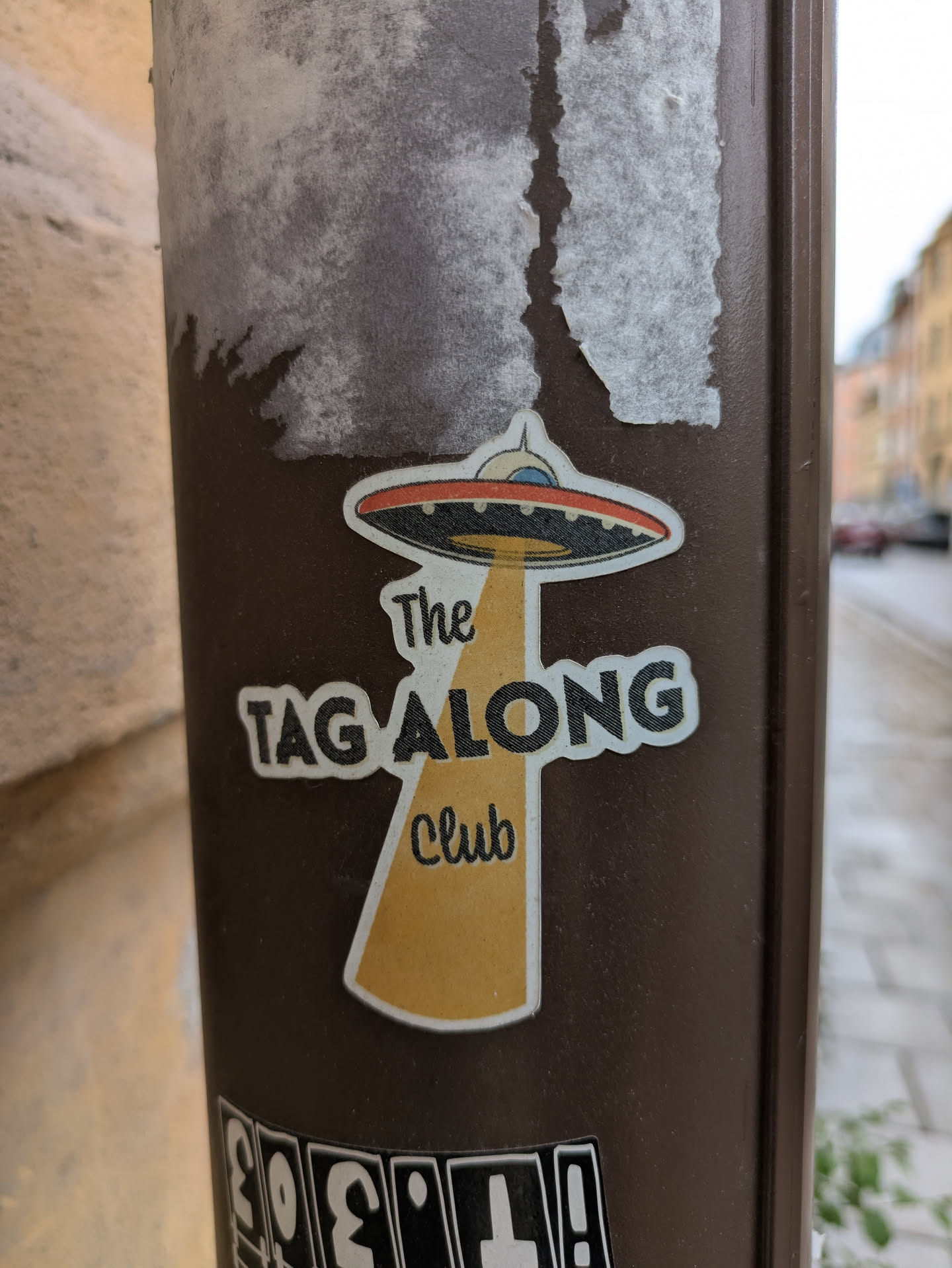 This sticker features an illustration of a flying saucer with a beam of light shining down from it. Underneath the beam of light are the words "The Tag Along Club". The sticker is on a brown pole.