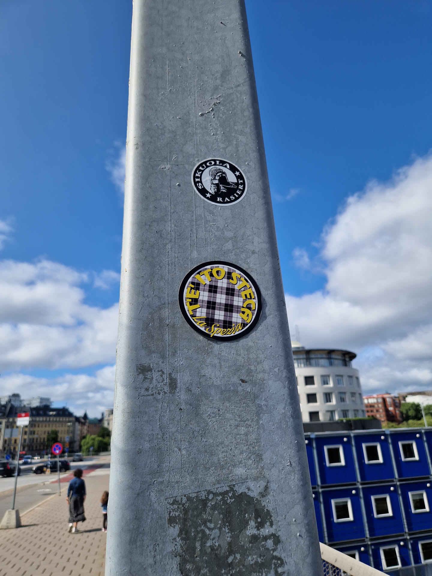 A round sticker with a yellow and black plaid pattern and the text "TIE DYE" is affixed to a metal pole outdoors. The sticker is the central focus of the image, with blurry buildings and a sky in the background.  There is a second sticker above, which is not in focus.