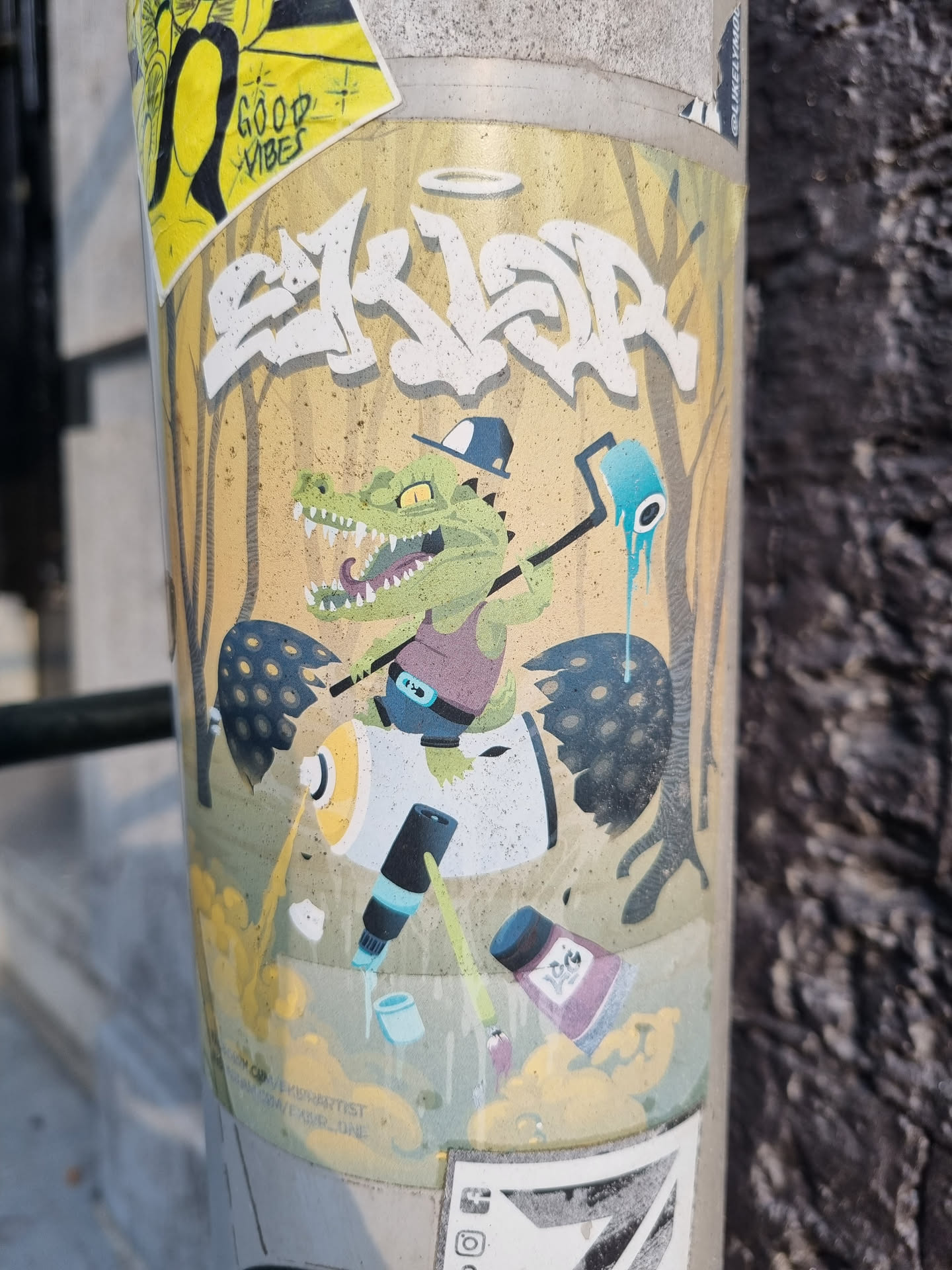 This sticker features a cartoon crocodile wearing a hat and holding a paint roller, surrounded by graffiti elements and paint cans. The style is playful and colorful. The text at the top says "SKLDR" in graffiti style and there is a small halo above the word. At the bottom there is small text that is hard to read.  There are no social media links or QR codes visible in the image.