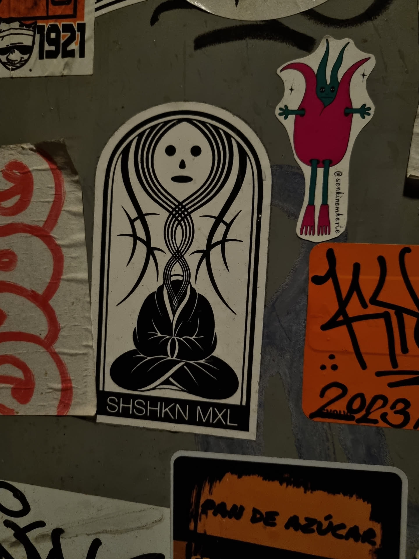 A black and white sticker featuring a figure in a meditative pose, surrounded by detailed linework. The sticker has a unique arched shape and includes the text 'SHSHKN MXL' at the bottom. 