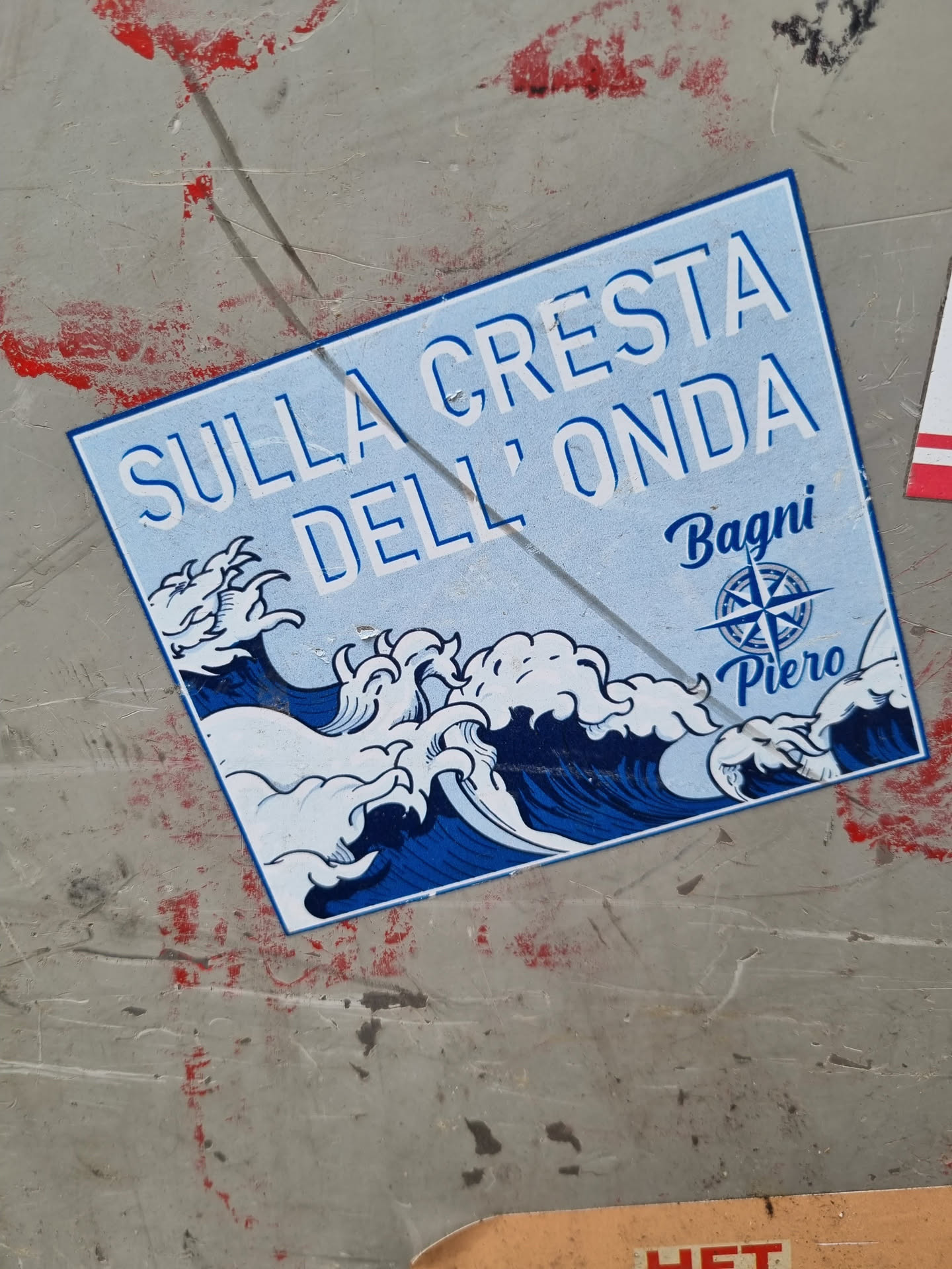 A blue and white sticker depicting stylized ocean waves, with the text "SULLA CRESTA DELL'ONDA" (On the Crest of the Wave) above a compass rose and "Bagni Piero" below it. The sticker has a slightly distressed look, fitting for its placement on a weathered surface.
