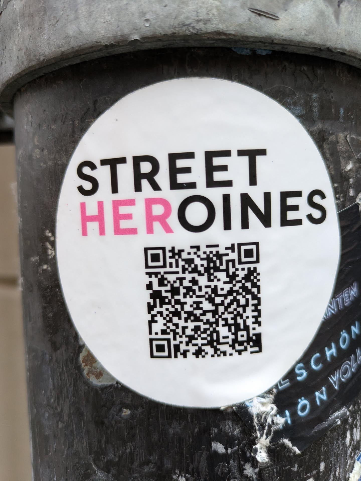 Round white sticker with the text STREET HEROINES in black and pink. It contains a QR code in the lower half. The sticker is affixed to a dark gray metallic surface.