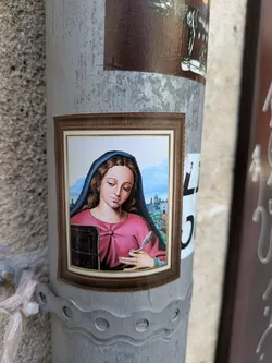 A framed sticker adhered to a pole features a portrait of a woman with brown hair and a dark blue veil, dressed in a pink top. She is holding a quill in her right hand and a closed book in her left hand. In the background is a landscape with a castle on the hill.
