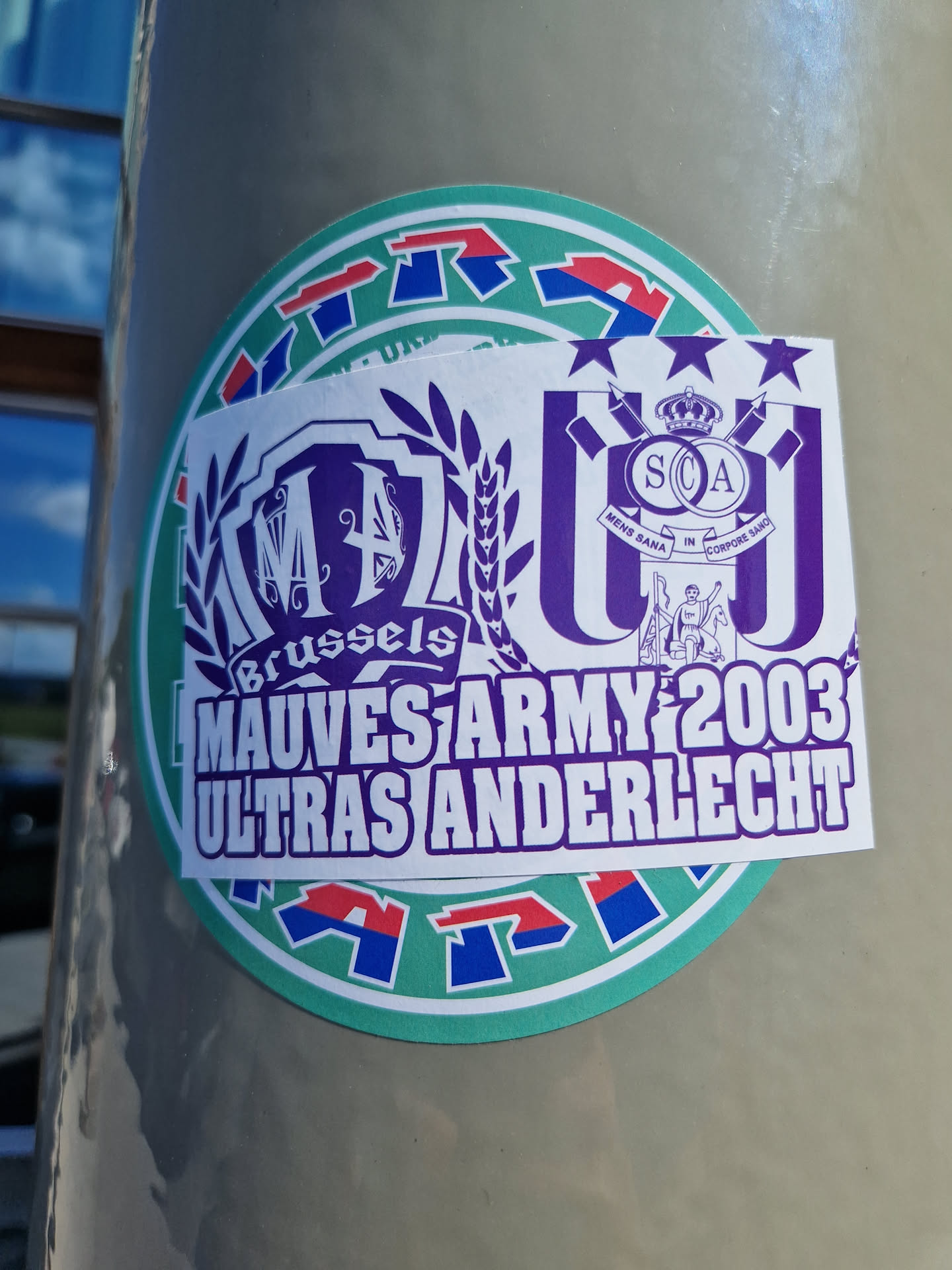 A sticker featuring the logo of Mauves Army 2003, the ultras group of Anderlecht. The sticker also includes the logos of two other groups. The main colors are purple and white.