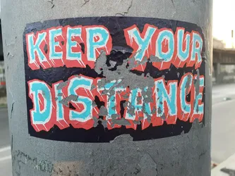 A worn sticker with the phrase "KEEP YOUR DISTANCE" in stylized, bold lettering. The colors are predominantly teal and coral on a dark background.  The sticker shows signs of wear and tear, with some peeling and damage to the surface.
