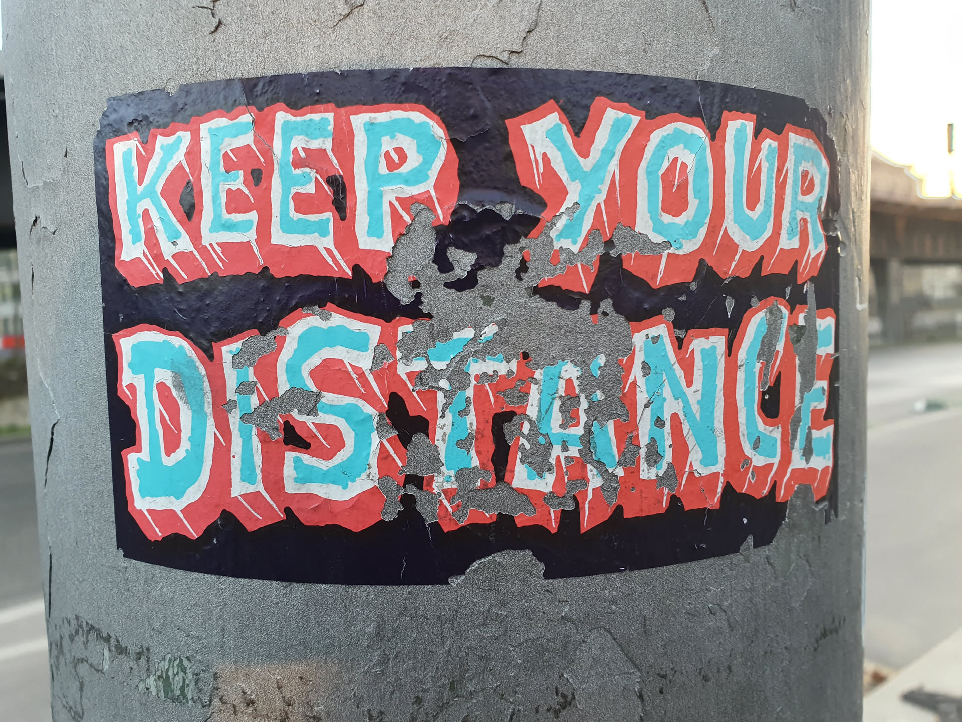 A worn sticker with the phrase "KEEP YOUR DISTANCE" in stylized, bold lettering. The colors are predominantly teal and coral on a dark background.  The sticker shows signs of wear and tear, with some peeling and damage to the surface.