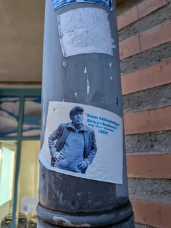 A rectangular off-white sticker affixed to a grey metal pillar. It features a blue-toned photograph of Swedish author and artist Stig 'Slas' Claesson wearing a flat cap and jacket with a cigarette in his mouth, standing with hands on his hips. Next to him, bold blue text in Swedish quotes his sentiment against Djurgården ('Dom blårandiga! Dra åt helvete.') followed by his name and the year '1962'.