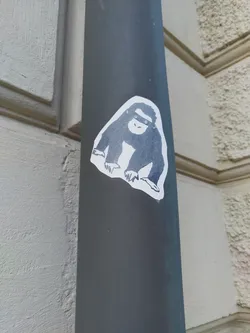 A sticker featuring a simple, grayscale drawing of a monkey. The monkey is depicted in a seated position, with its hands resting on its knees. The sticker is affixed to a dark gray pole.