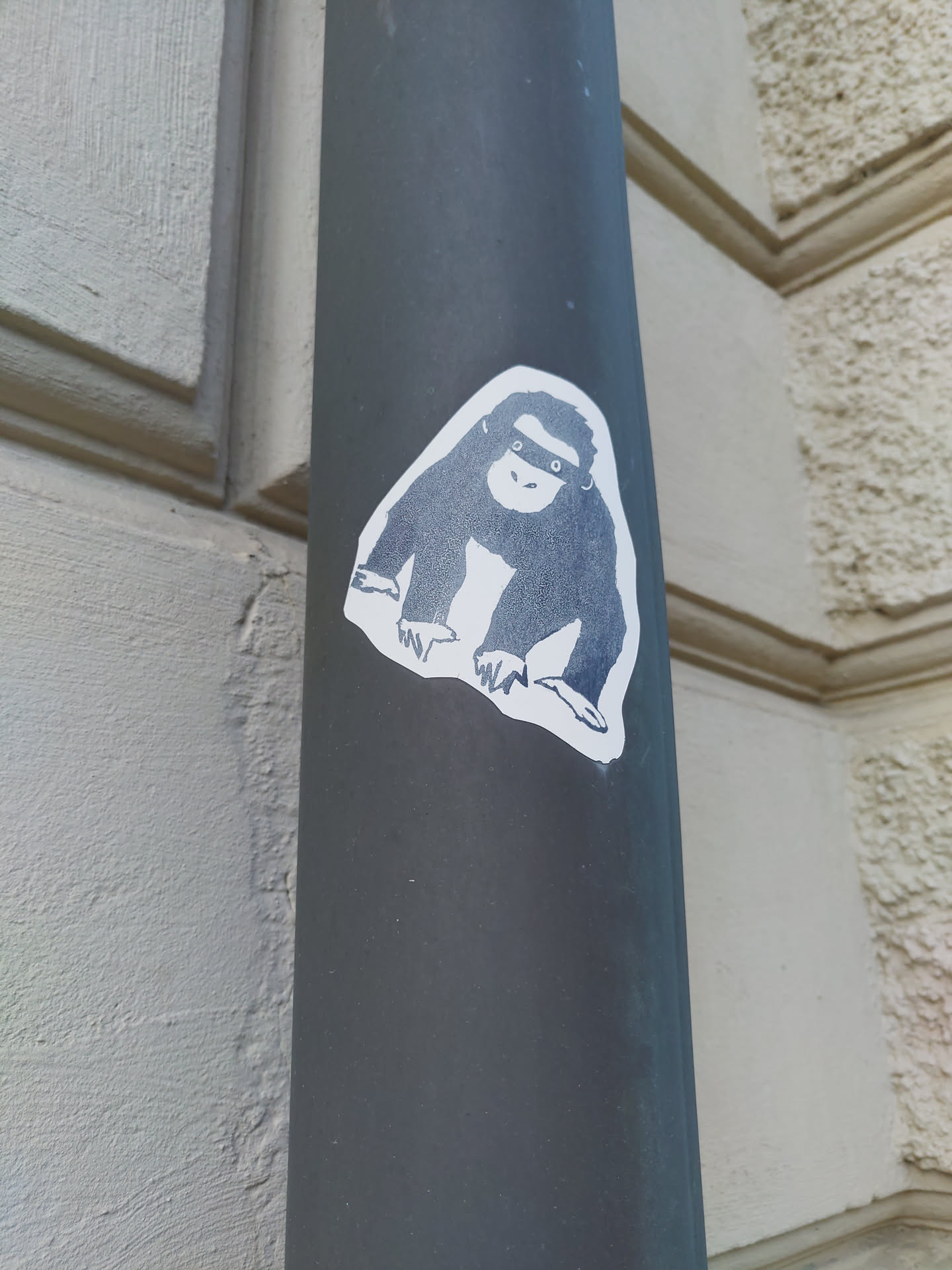 A sticker featuring a simple, grayscale drawing of a monkey. The monkey is depicted in a seated position, with its hands resting on its knees. The sticker is affixed to a dark gray pole.