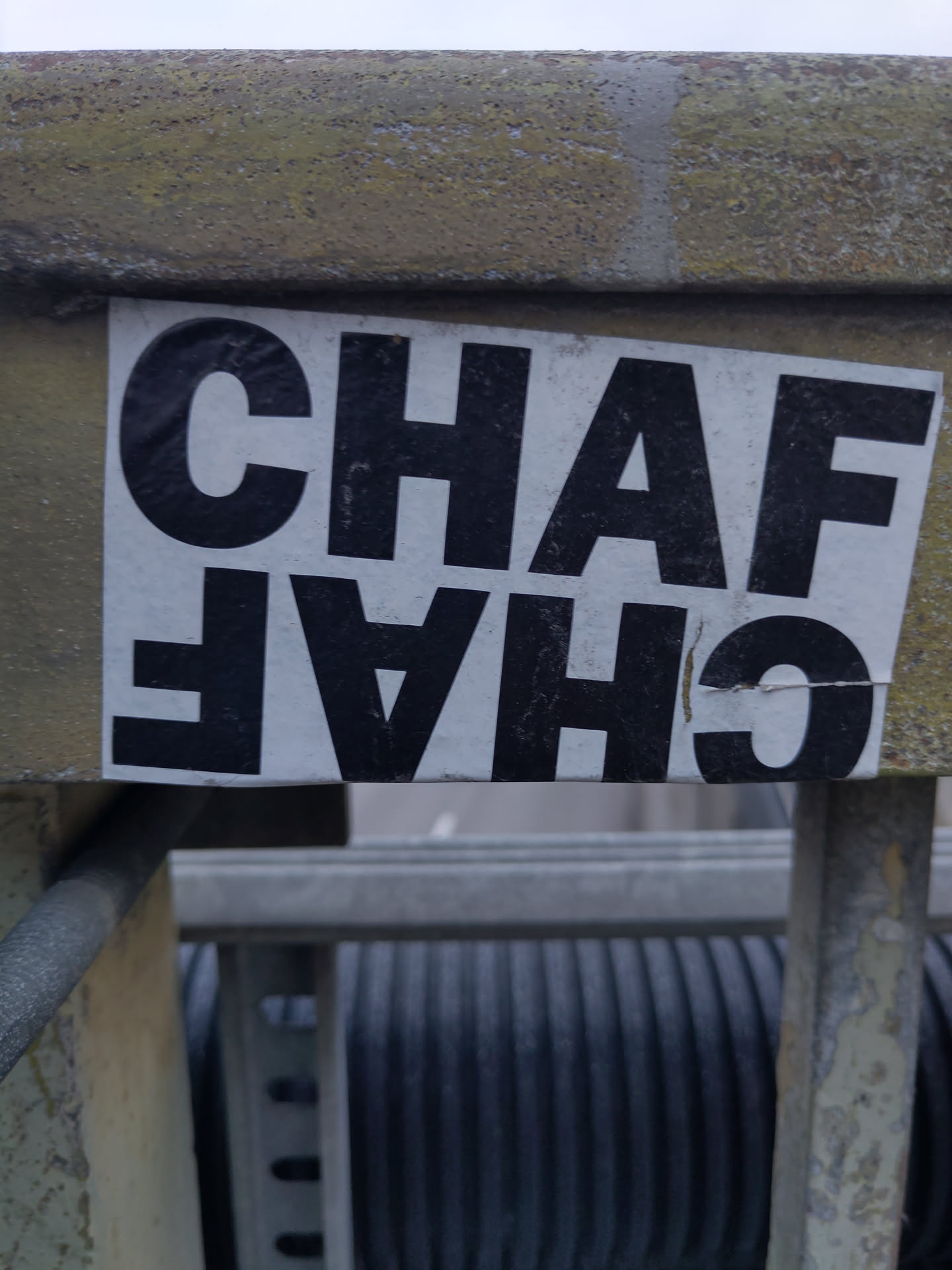 A black and white sticker with the text "CHAFA" written in a bold, sans-serif font. The sticker is affixed to a weathered, light-grey metal surface. The text is vertically inverted. 