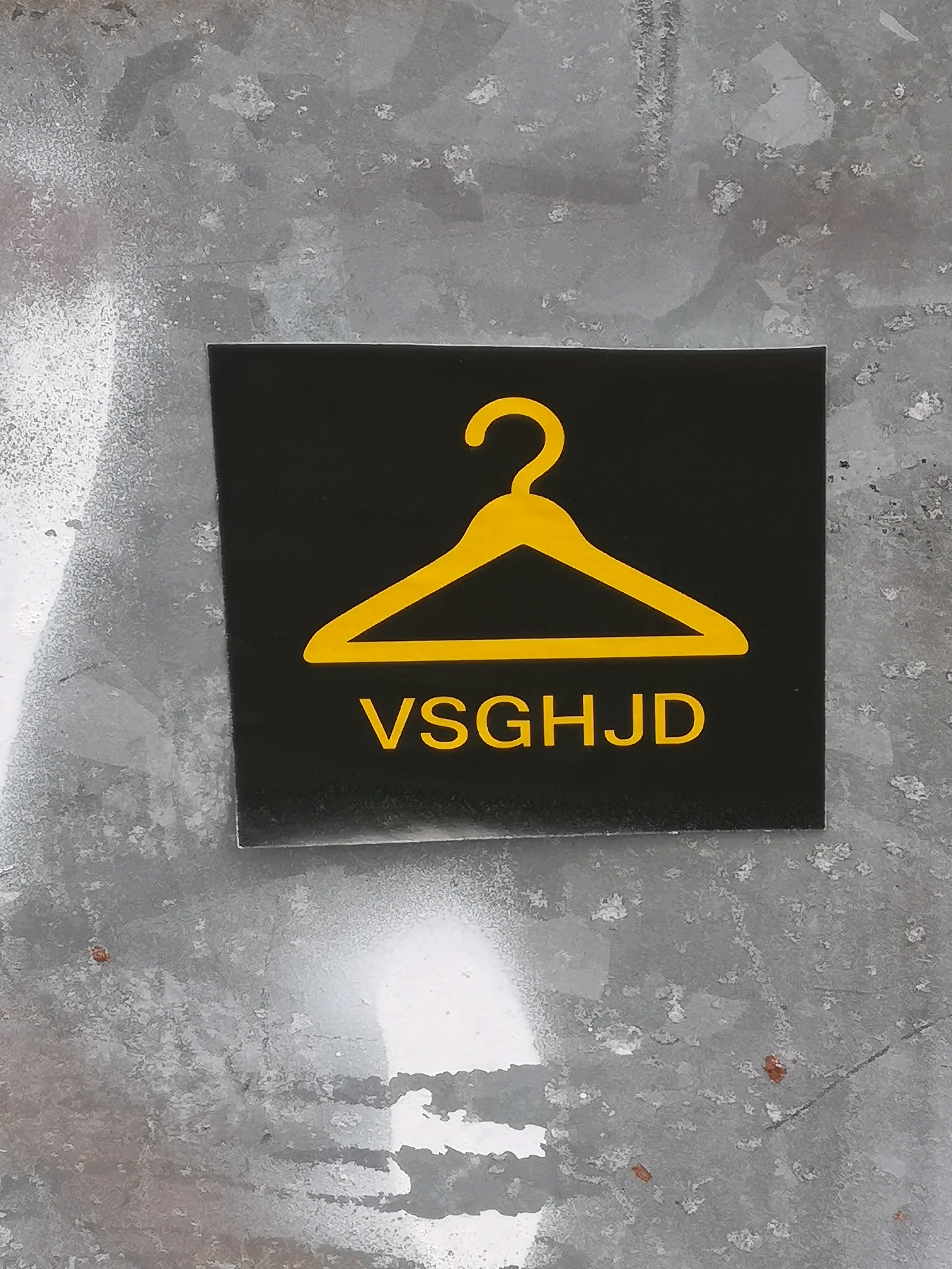 A rectangular sticker with a solid black background. Centered in the upper half is a bright yellow, stylized outline of a clothing hanger. Below the hanger, the capital letters "VSGHJD" are displayed in a bold, sans-serif font, also in bright yellow. The sticker has a clean, high-contrast design against a textured, grey wall.
