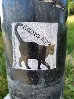 A sticker featuring a black cat silhouette with the text 'Adora Eye' and a Facebook link. The sticker is on a dark-colored pole.