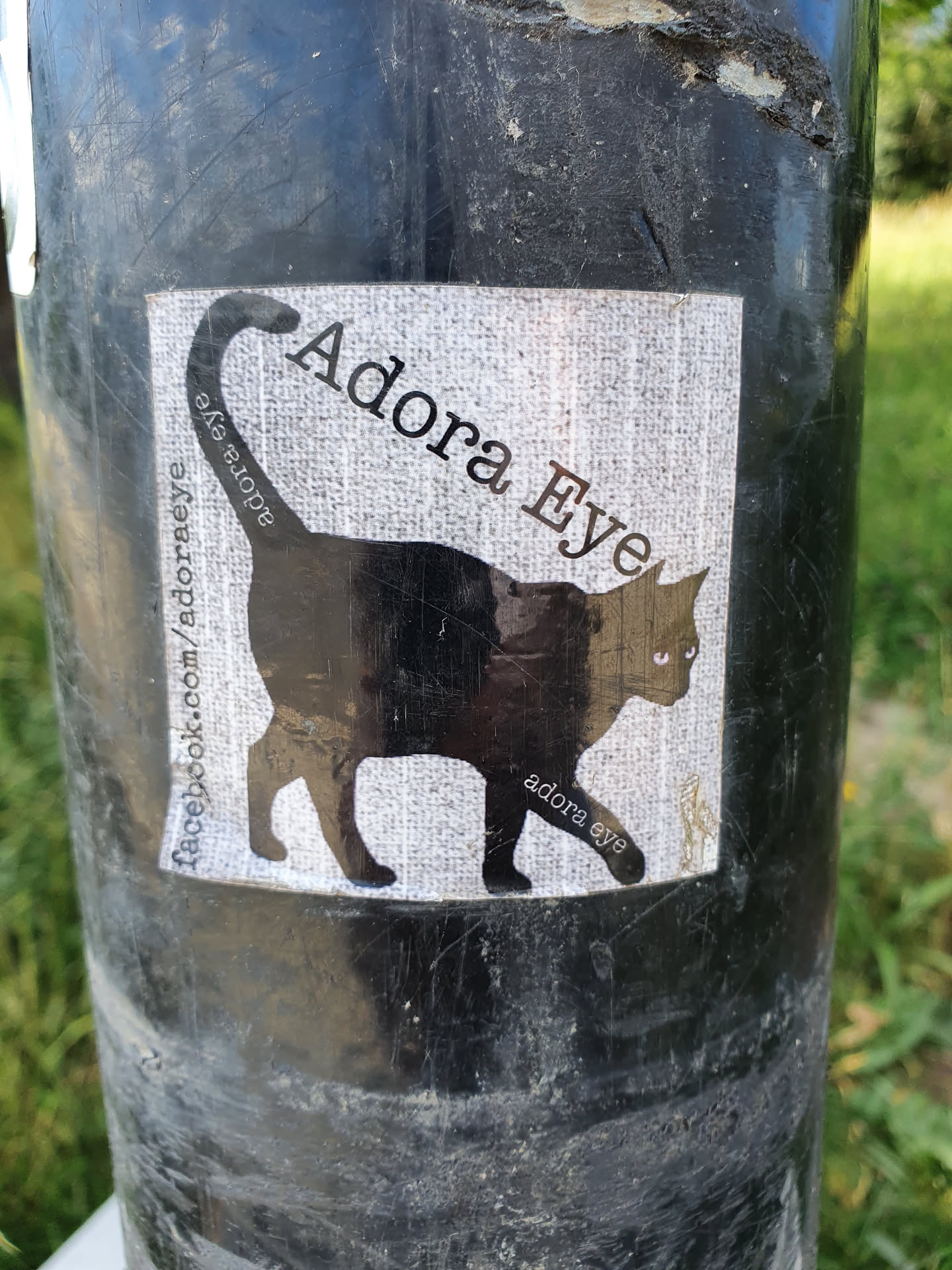 A sticker featuring a black cat silhouette with the text 'Adora Eye' and a Facebook link. The sticker is on a dark-colored pole.