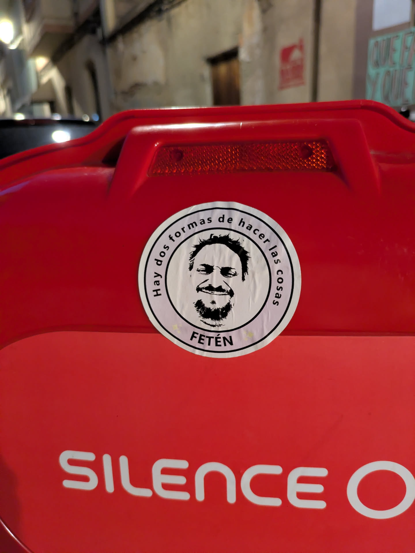 This circular sticker is placed on a red surface, likely a vehicle or container. The sticker features a portrait of a man with a beard, and the text "Hay dos formas de hacer las cosas" and "FETÉN" can be read on the sticker.