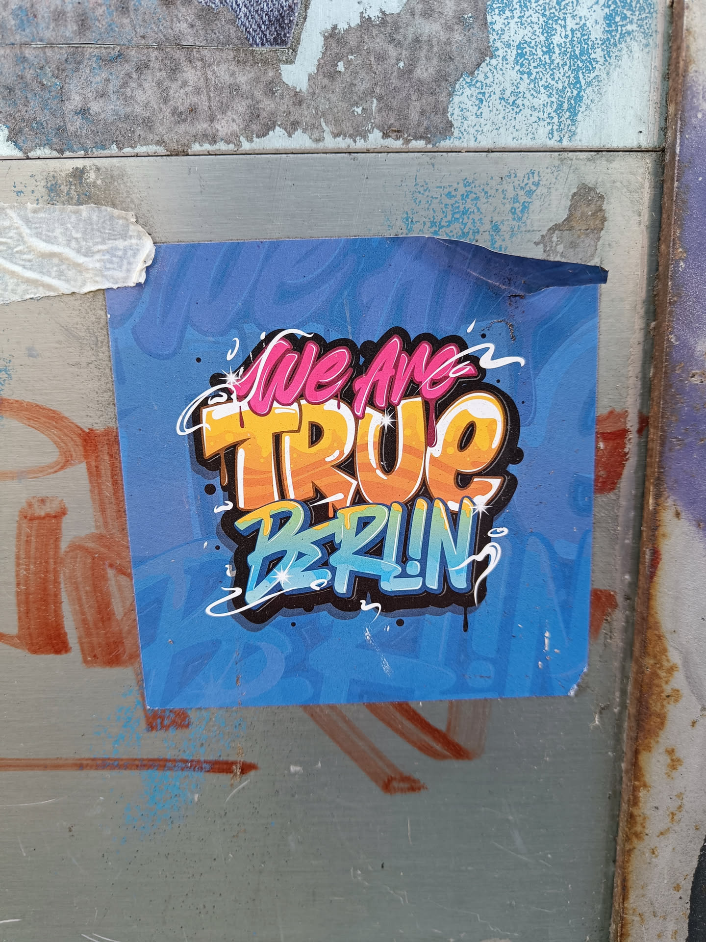 A blue sticker with graffiti-style text reading "We Are True Berlin". The text is in a mix of pink, orange, and blue colors, with a slightly worn appearance. It's affixed to a weathered surface with visible scratches and other graffiti.