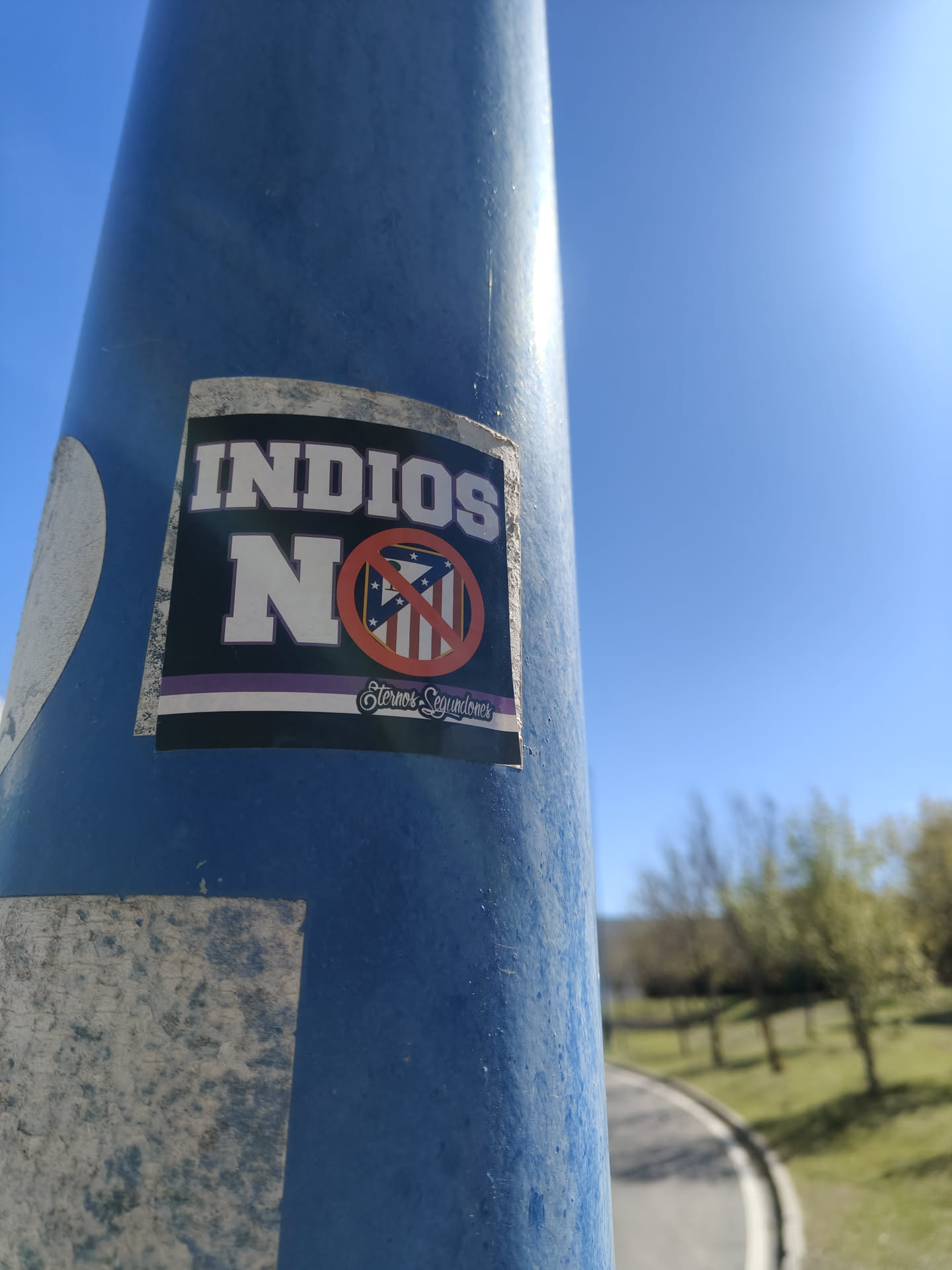 A rectangular sticker with a dark blue/black background, featuring prominent white text. At the top, 'INDIOS' is written in large, bold white capital letters. Below it, a large white 'N' is followed by a red prohibition circle with a diagonal line, overlaying a shield-shaped emblem. This emblem resembles the Atlético Madrid club crest, with vertical red and white stripes, a blue section with white stars, and a bear and tree motif. Below this central design, there's a thin white stripe, a thin purple stripe, and another thin white stripe. At the very bottom, in a stylized white script, the phrase 'Eternos Segundones' is written. The sticker shows some wear and tear, particularly around its edges, and is affixed to a blue pole.