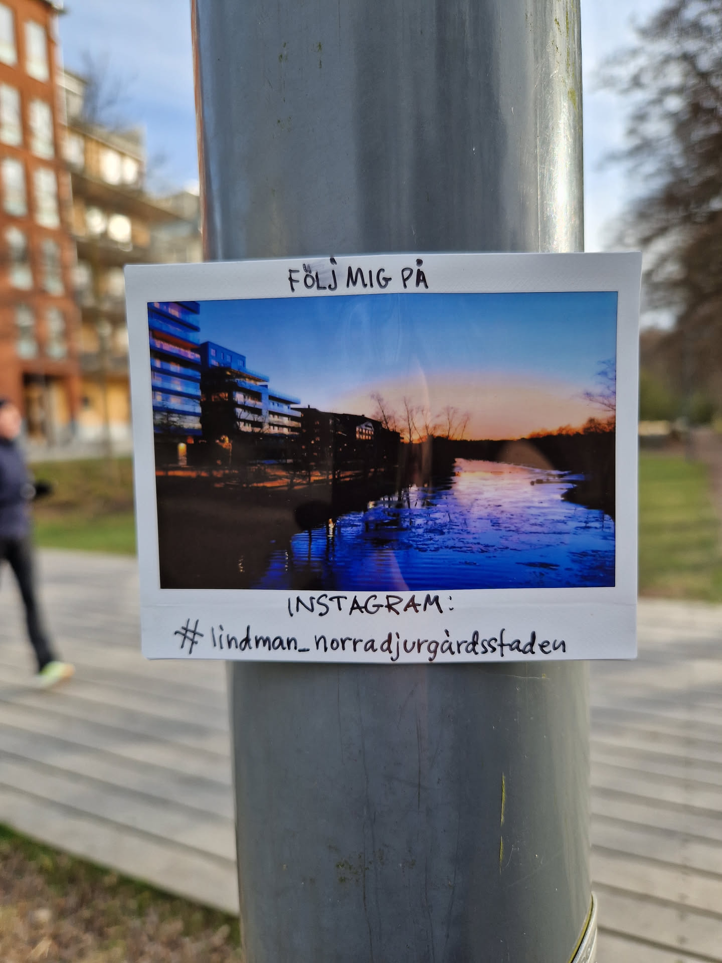 A Polaroid-style photo sticker featuring a sunset view over a river and modern buildings.  The sticker is affixed to a pole and includes a handwritten Instagram handle.