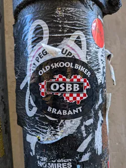 A circular black sticker placed on a worn, textured pole over older peeling stickers. The top of the sticker reads 'OLD SKOOL BIKER' in an arched, bold white font. In the center is a red-and-white checkered silhouette representing the Dutch province of North Brabant, overlaid with a black rectangle outlined in white containing the bold acronym 'OSBB'. At the bottom, curved along the lower perimeter, the word 'BRABANT' is printed in white capital letters.