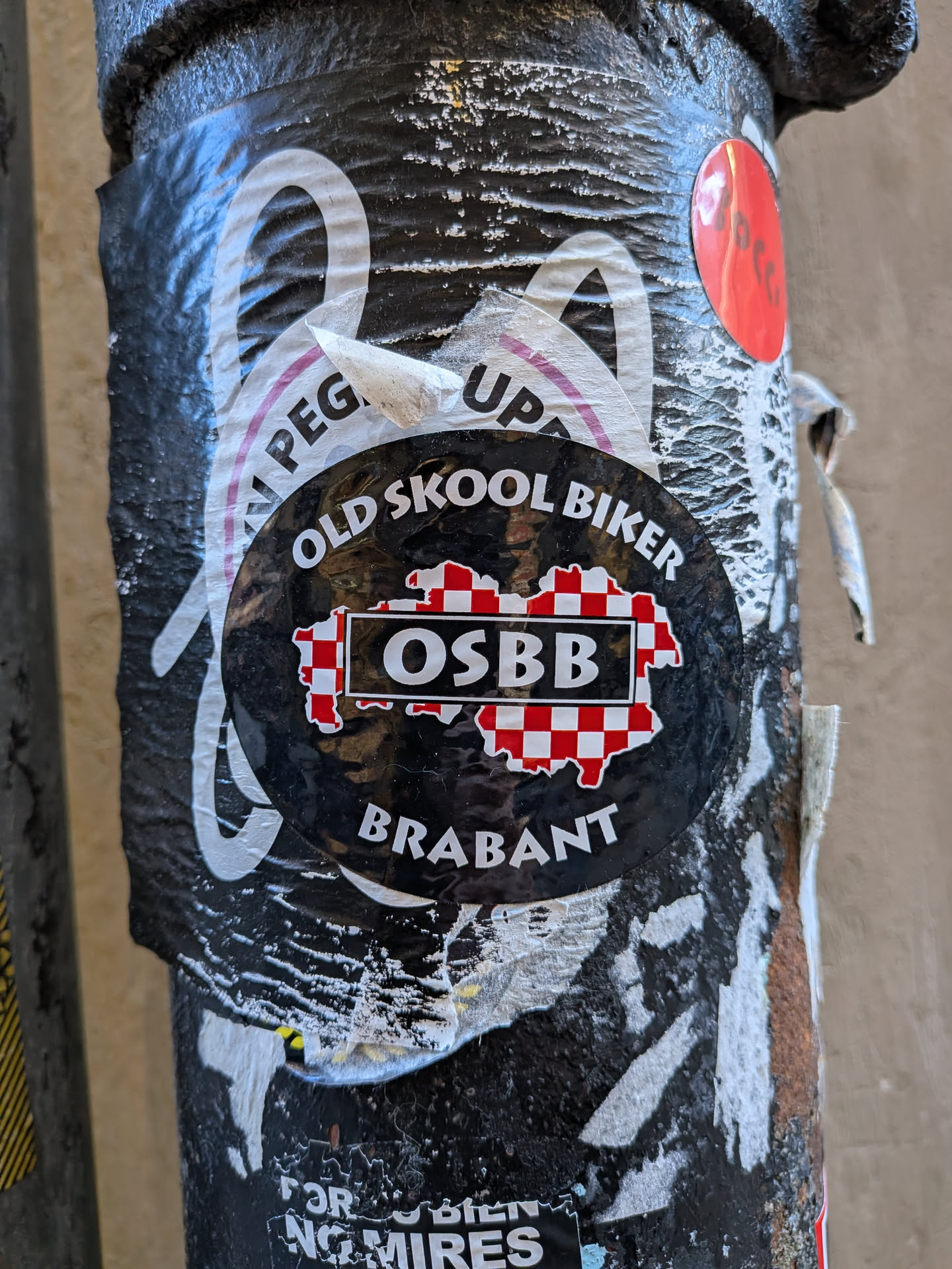 A circular black sticker placed on a worn, textured pole over older peeling stickers. The top of the sticker reads 'OLD SKOOL BIKER' in an arched, bold white font. In the center is a red-and-white checkered silhouette representing the Dutch province of North Brabant, overlaid with a black rectangle outlined in white containing the bold acronym 'OSBB'. At the bottom, curved along the lower perimeter, the word 'BRABANT' is printed in white capital letters.